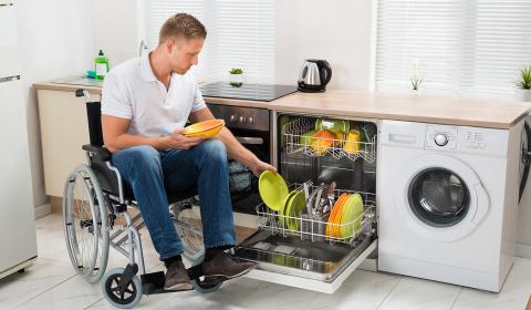 Un jeune homme en fauteuil roulant, de profil dans une cuisine, charge le bac d'un lave-vaisselle